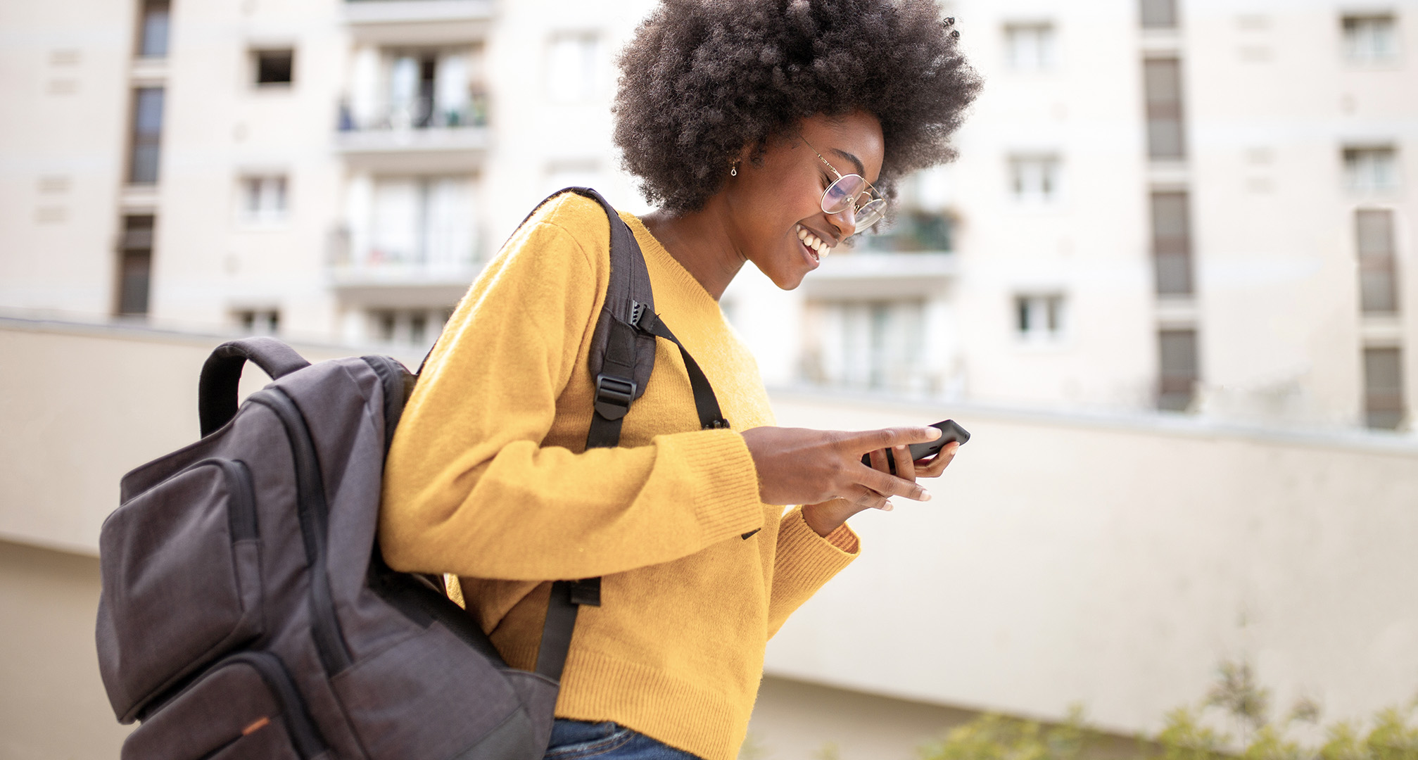 person with yellow sweater, wire-rimmed glasses, and grey backpack smiling at phone screen outside