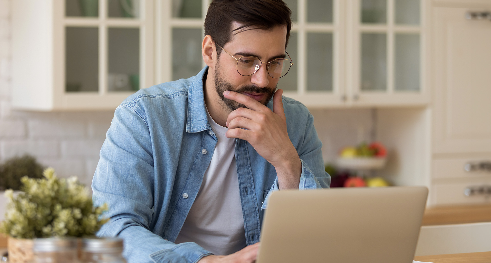 person with glasses sitting in kitchen looking at laptop